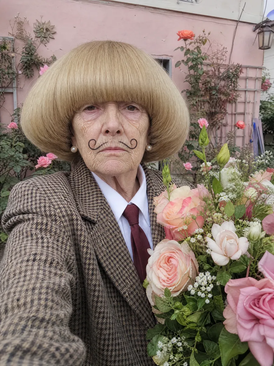Odette, a Jean Phil-style AI character by Starrd: a grandmother with a huge blond bob and a curled moustache drawn on in eyeliner, holding a bouquet